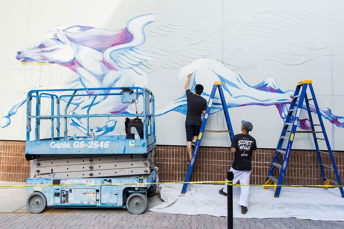 Part of a white horse is painted on a white background. There is a blue lift in front of it with two blue ladders. Artist Boy Kong is seen from behind painting in the side of the horse while his brother watches next to him.