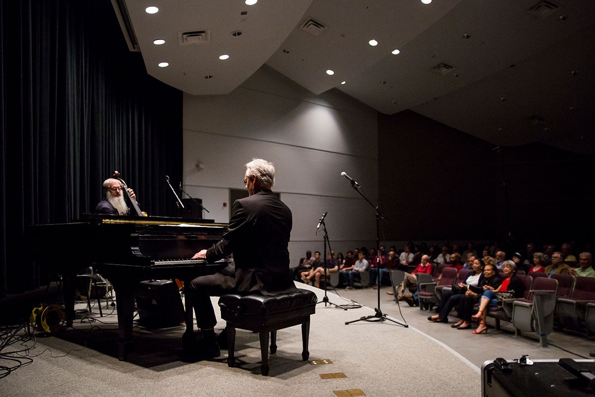 The man is seen playing the piano. His back is to the photographer. Facing him is a man playing an upright bass. The audience can be seen to the right.