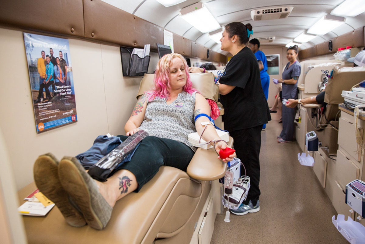 A woman with blond and pink hair is sitting in a beige lounge chair, with a red ball in her left hand, where blood is being drawn from her arm. A nurse looks over her.
