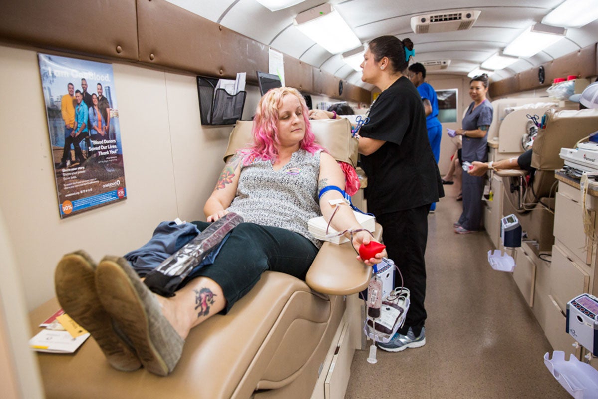 A woman with blond and pink hair is sitting in a beige lounge chair, with a red ball in her left hand, where blood is being drawn from her arm. A nurse looks over her. A woman with blond and pink hair is sitting in a beige lounge chair, with a red ball in her left hand, where blood is being drawn from her arm. A nurse looks over her.
