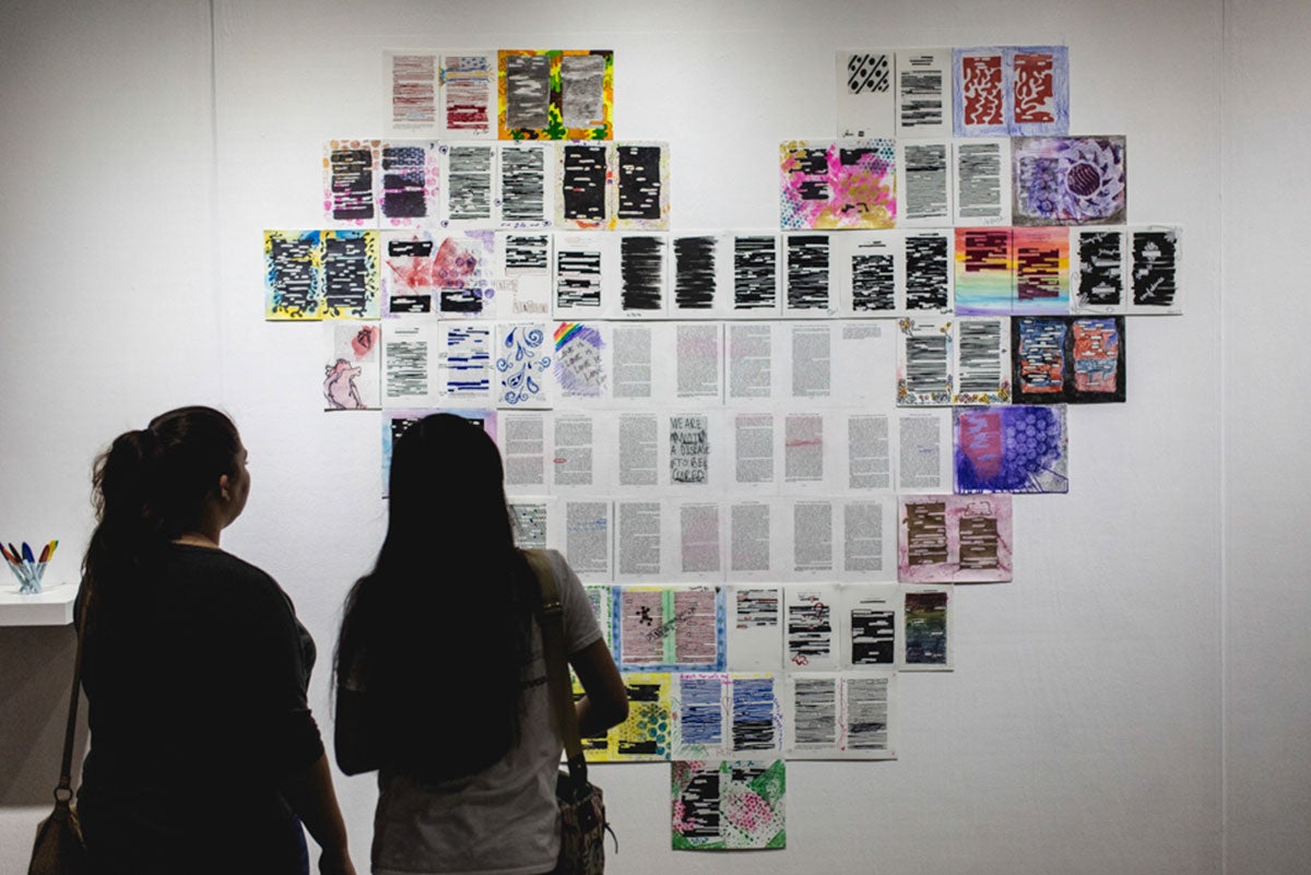 The silhouettes of two women are seen looking at a giant installation of pieces of paper torn from a book, colored, and shaped into a giant heart.