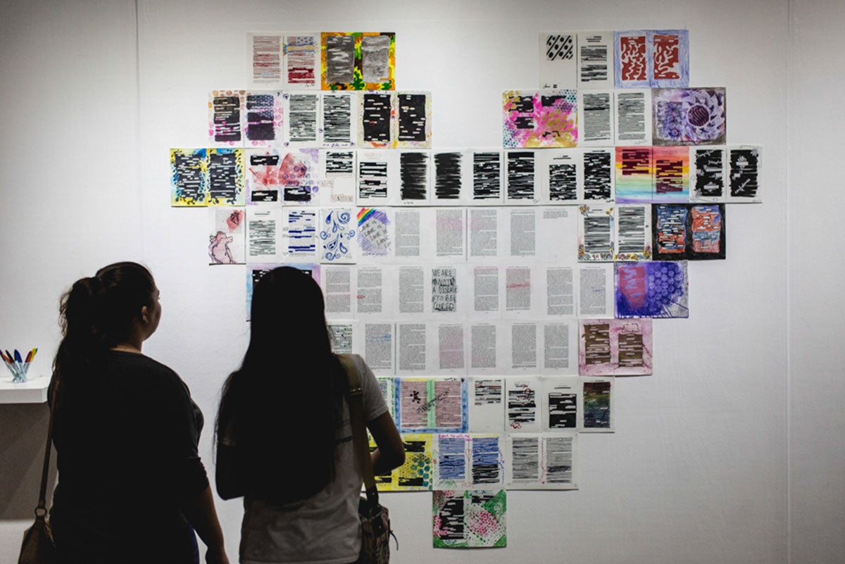The silhouettes of two women are seen looking at a giant installation of pieces of paper torn from a book, colored, and shaped into a giant heart. The silhouettes of two women are seen looking at a giant installation of pieces of paper torn from a book, colored, and shaped into a giant heart.