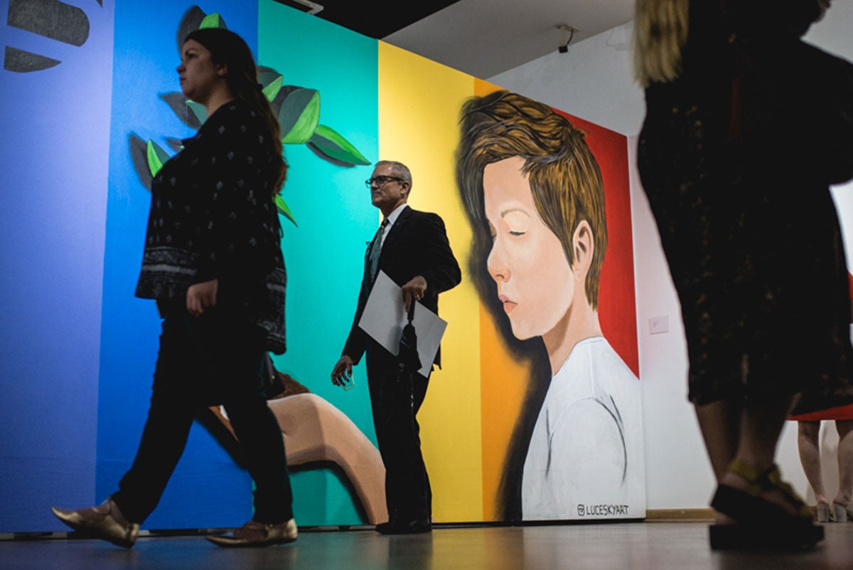People are seen walking in front of a large mural of an androgynous looking person with short brown hair wearing a white shirt on a rainbow-colored background.
