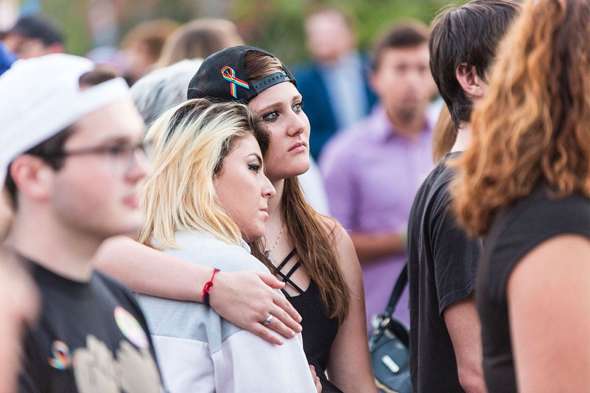 One woman in a black baseball hat turned backward with rainbow ribbon has her arm wrapped around another woman, who rests her head on the shoulder of the woman wearing the black hat.