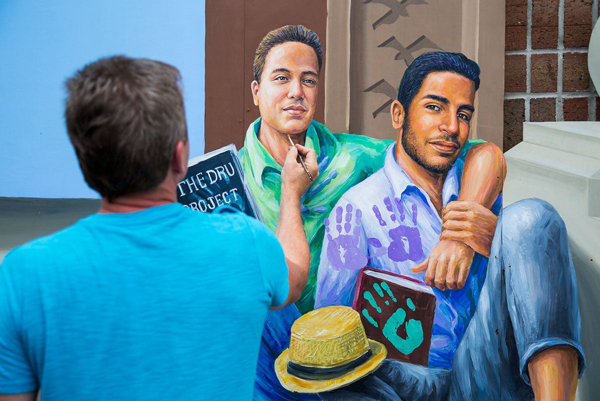 A man in a blue shirt is seen holding up a paintbrush to the chin of a painting of two men sitting together, one with his arm around the other.