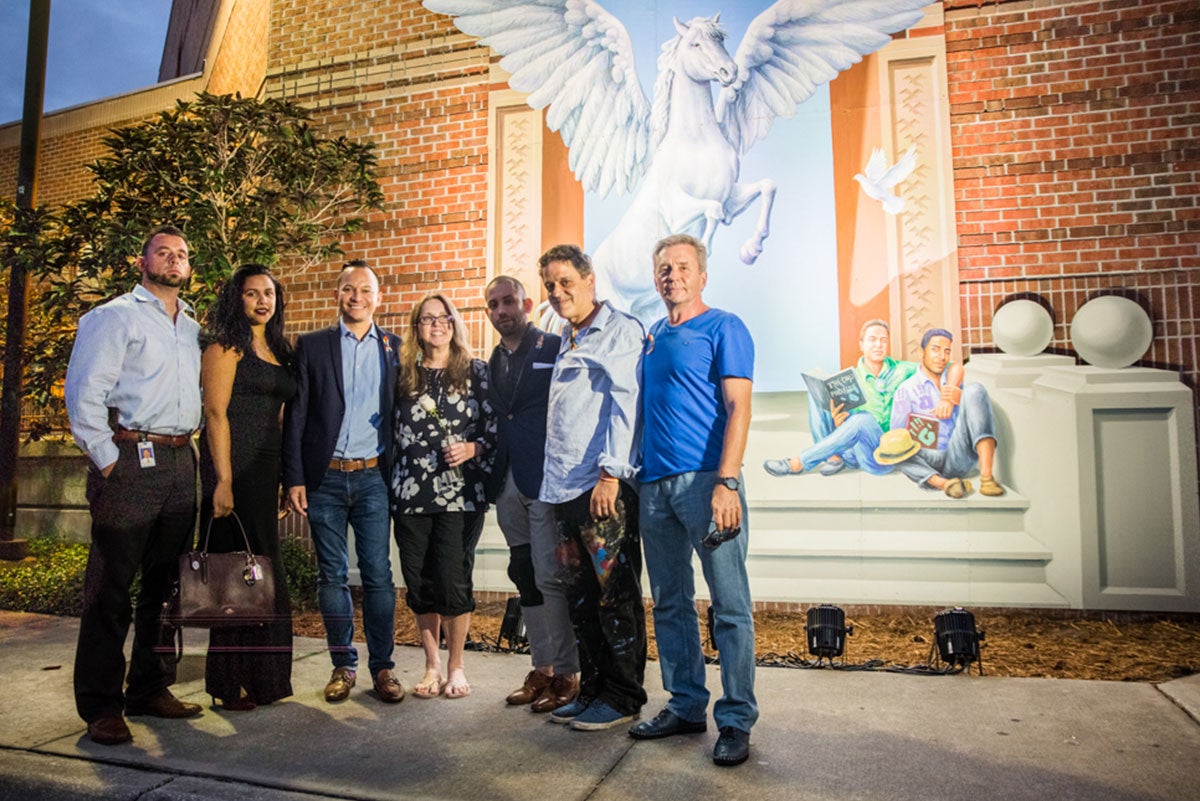 Five men and two women stand together facing the camera in front of the mural, which is lit up on a wall behind them. You can see a flying white horse rearing up and two men sitting on steps on the mural.