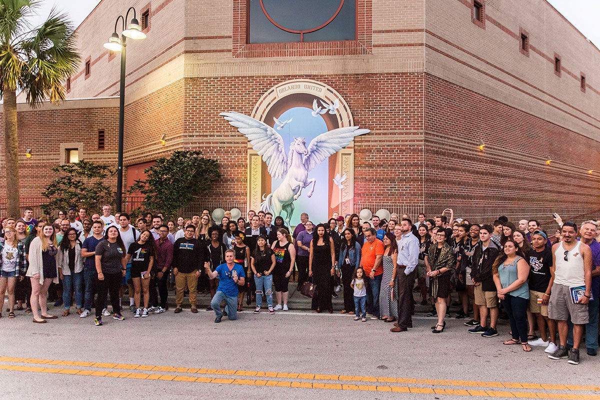 A large group of people stand facing the camera in front of the mural. You can see the white, winged horse rearing in the mural and the part of the Student Union.