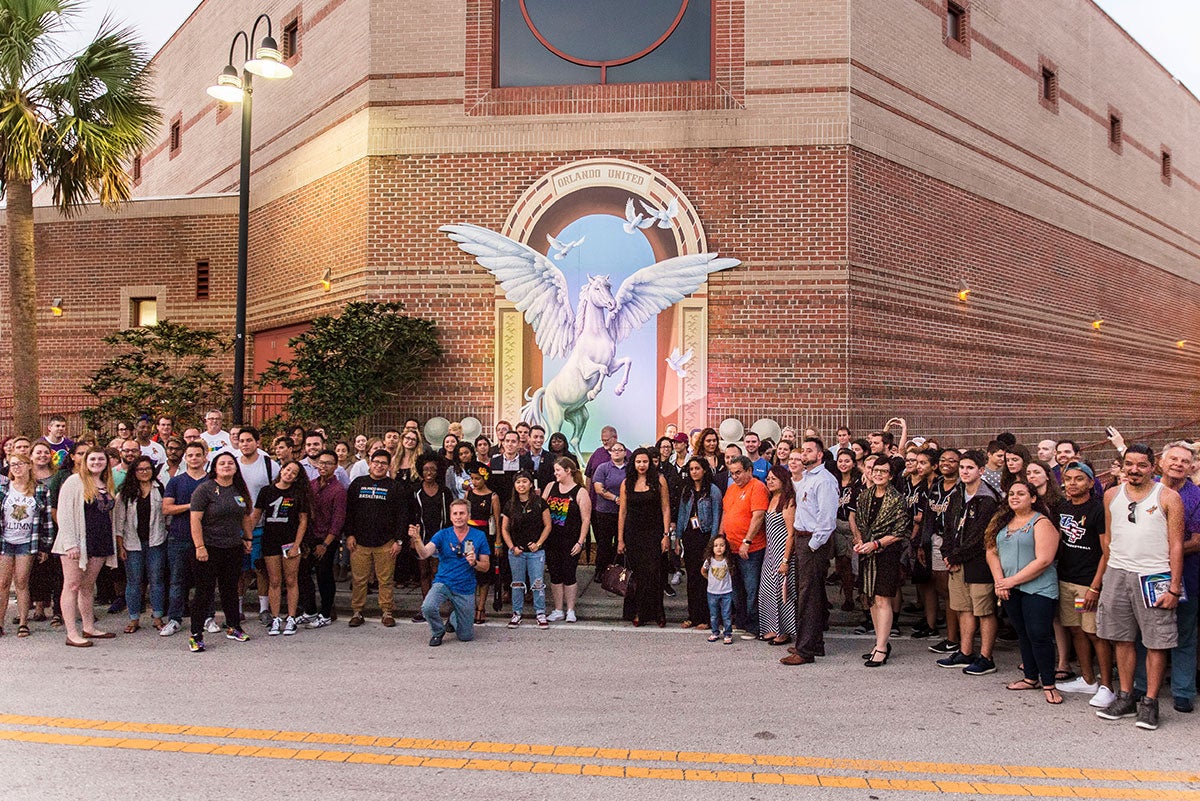A large group of people stand facing the camera in front of the mural. You can see the white, winged horse rearing in the mural and the part of the Student Union. A large group of people stand facing the camera in front of the mural. You can see the white, winged horse rearing in the mural and the part of the Student Union.