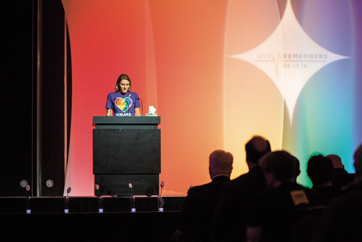 A woman wearing a purple Orlando United shirt stands in front of a podium. Behind her is a screen with a giant rainbow. A woman wearing a purple Orlando United shirt stands in front of a podium. Behind her is a screen with a giant rainbow.