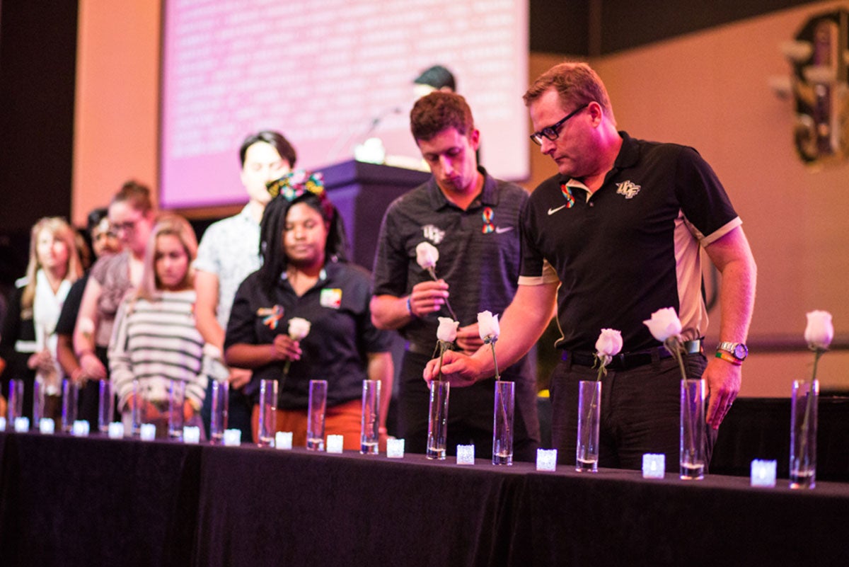 Eight people are seen holding white roses while standing in front of a row of clear vases on a black table. The person in focus in the right is placing his rose in one of the vases.