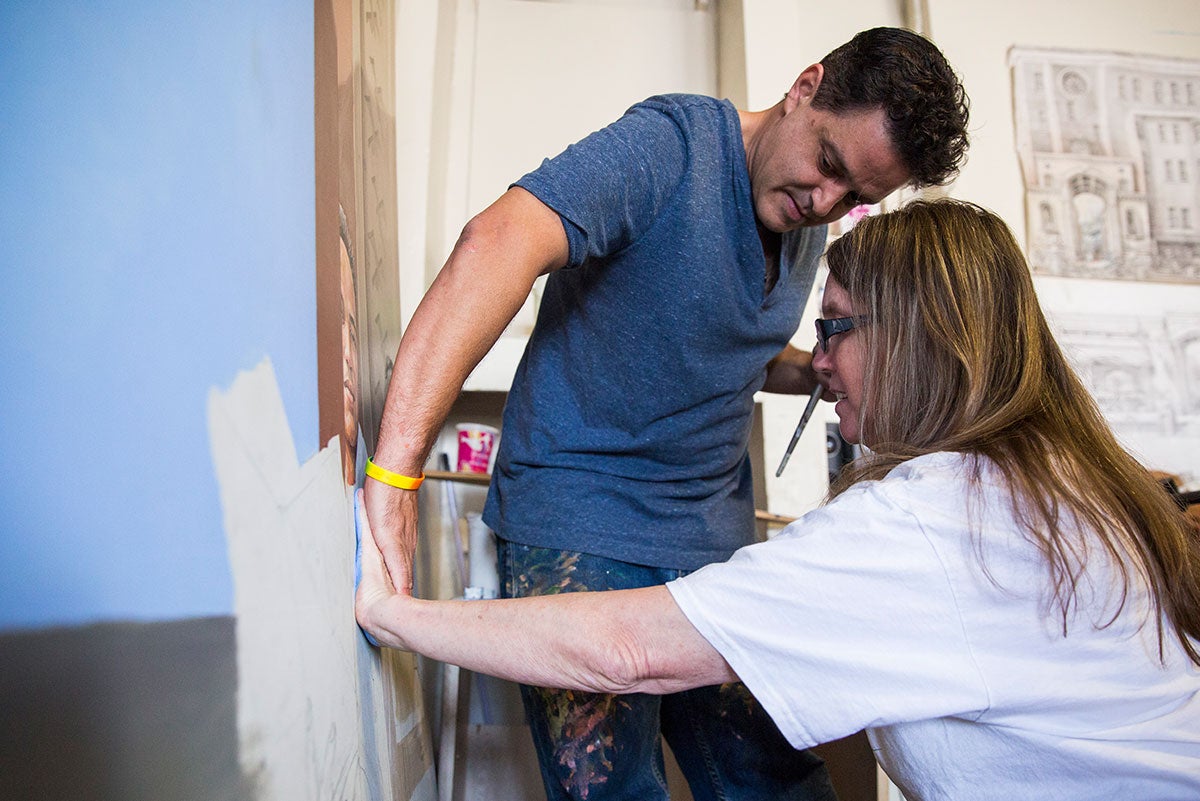 A woman in a white shirt holds her hand against a canvas while a man in a blue shirt puts his hand on top of hers, helping to soak the paint.
