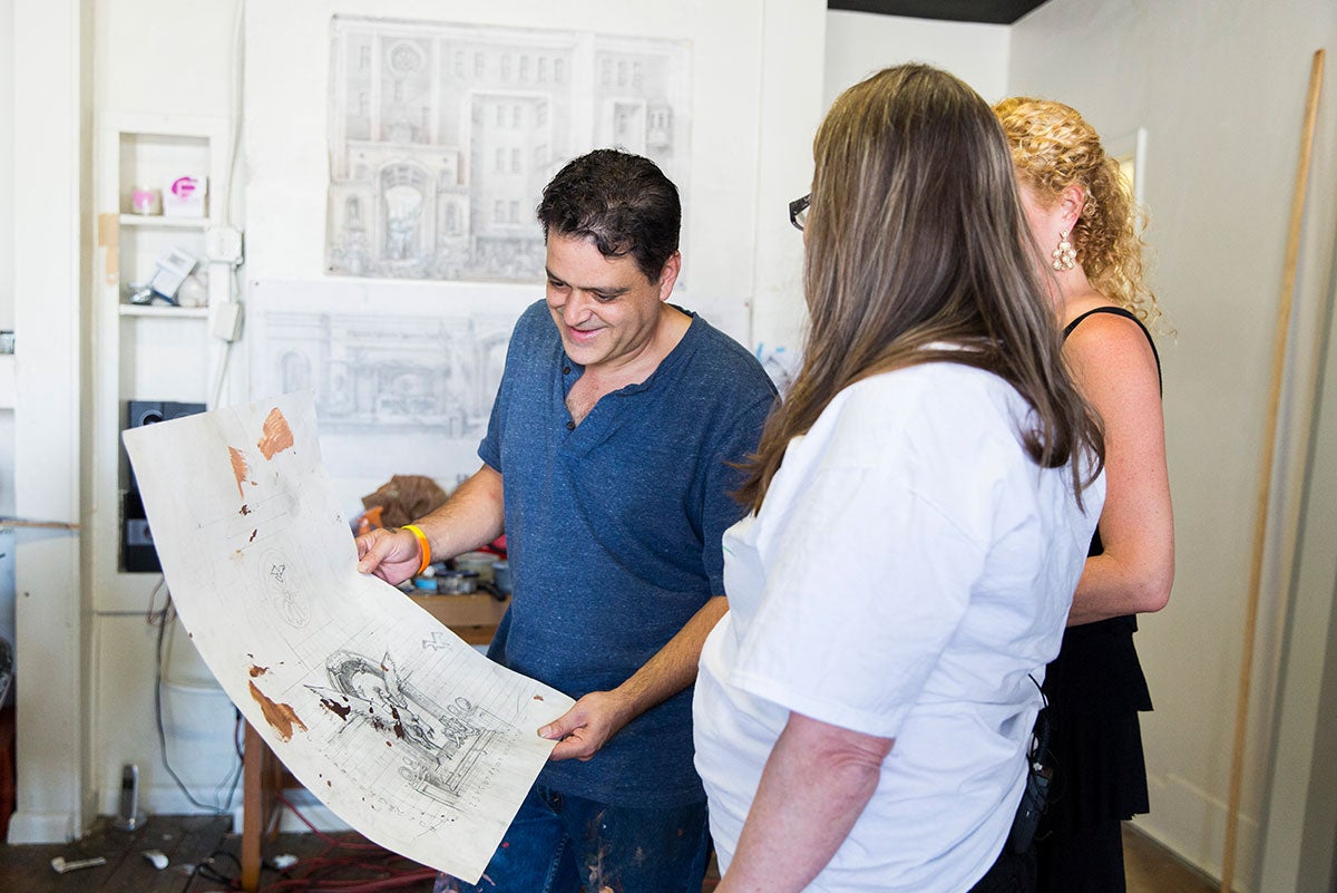 A man in a blue shirt is holding a large piece of paper with a sketch and paint stains as two women look on. A man in a blue shirt is holding a large piece of paper with a sketch and paint stains as two women look on.