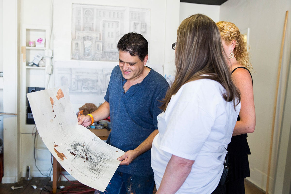 A man in a blue shirt is holding a large piece of paper with a sketch and paint stains as two women look on.