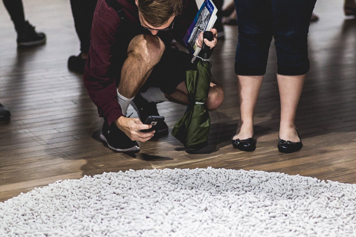 A man squats down, holding his cell phone over a large, round pile of small, white objects. The legs of woman standing next to him can be seen. She is wearing black ballet flats. A man squats down, holding his cell phone over a large, round pile of small, white objects. The legs of woman standing next to him can be seen. She is wearing black ballet flats.