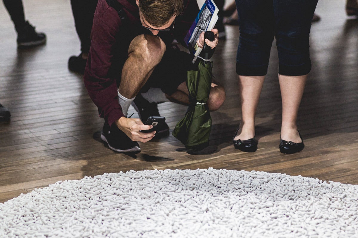 A man squats down, holding his cell phone over a large, round pile of small, white objects. The legs of woman standing next to him can be seen. She is wearing black ballet flats.