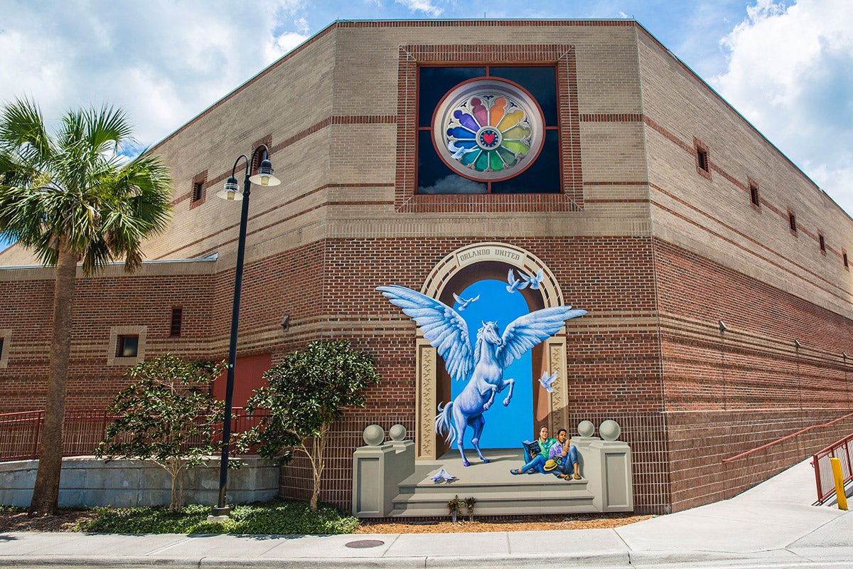 The Pulse mural at UCF is installed on a wall on the Student Union. On the brick wall is a tromp l'oeil painting of an archway, with a winged, white horse rearing up. It is standing at the top of a few concrete steps, which also has two men sitting together nearby holding books and each other. Above this is a round, rainbow-colored stained glass window with a heart in the middle and a single white dove in flight. 