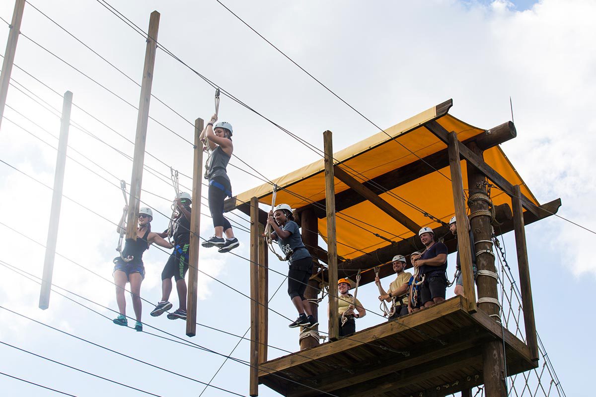 Four people wearing helmets try to cross wires suspended in the air.