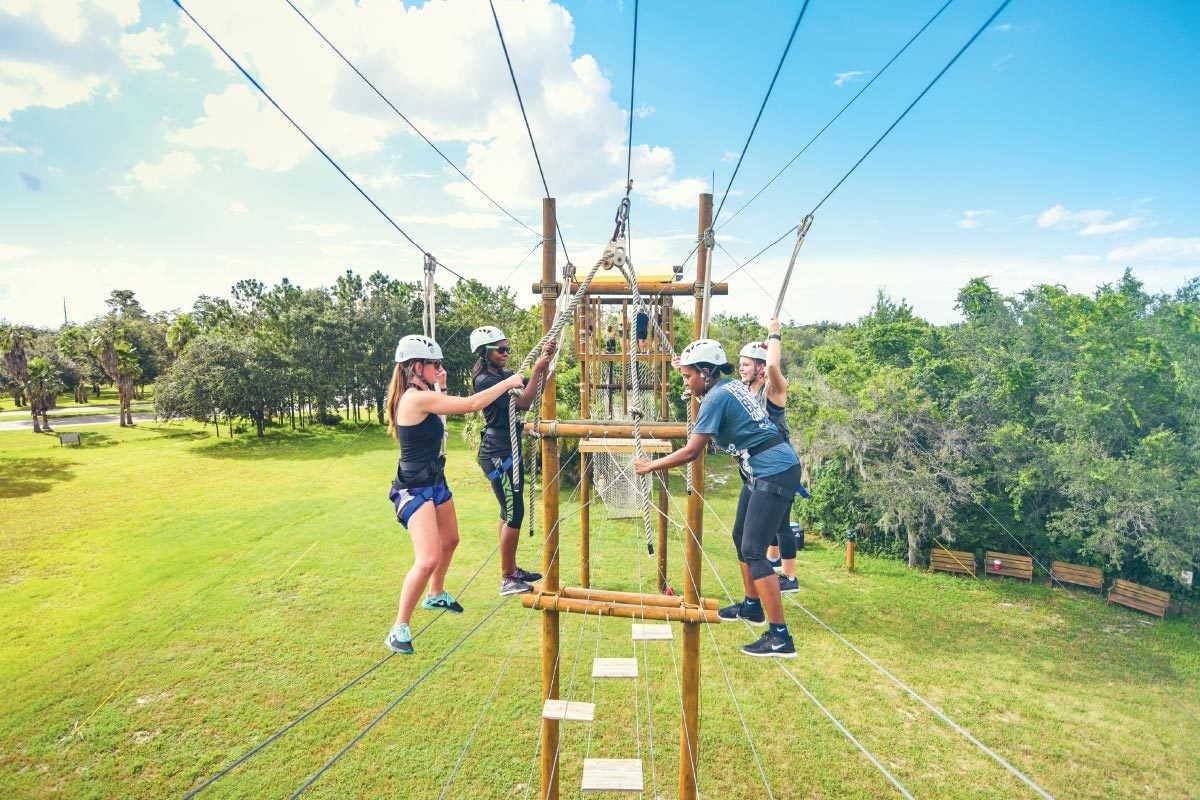 Four people in helmets hold onto ropes as they try to cross wires suspended in the air to complete a high-elements climbing course.