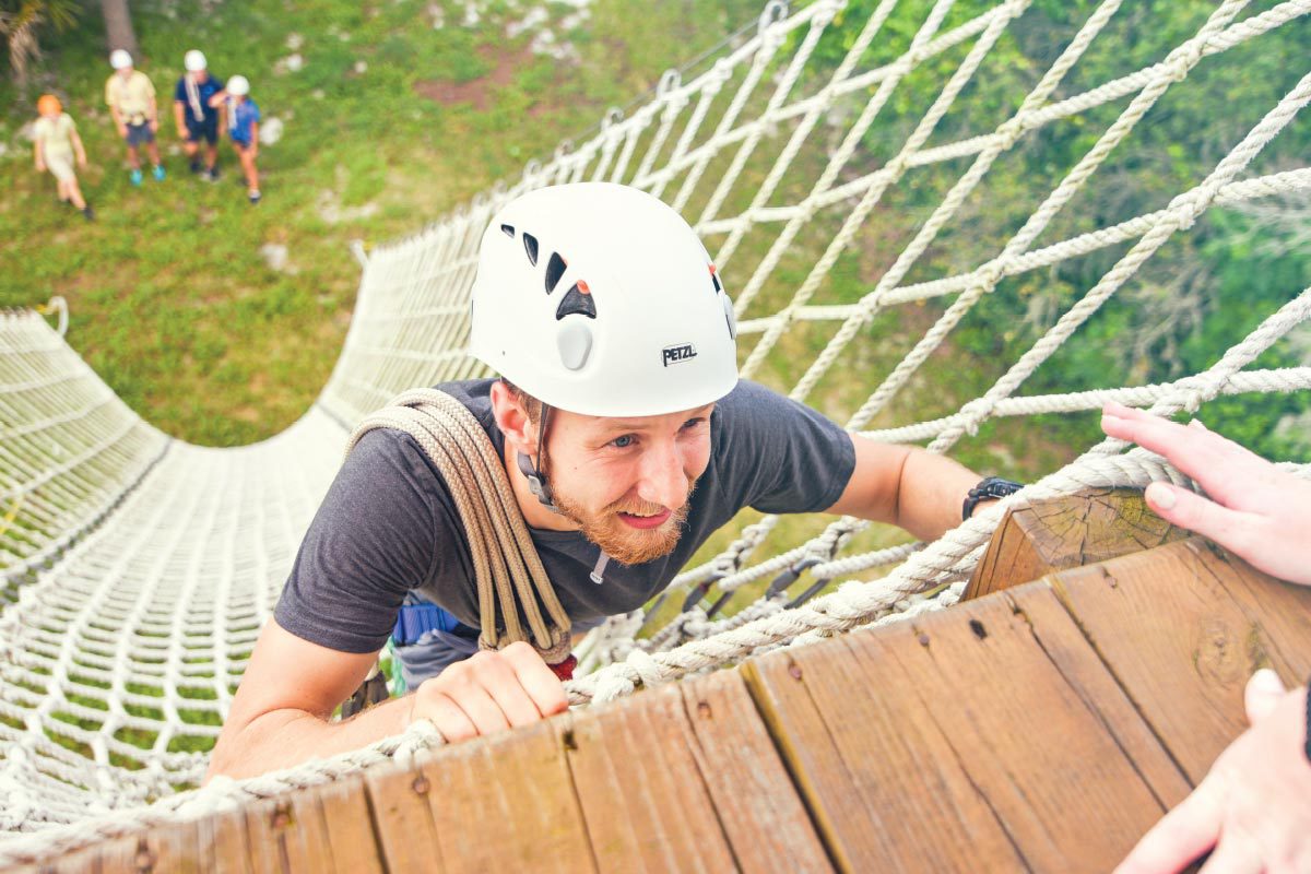 A man in a white helmet climbs a rope ladder to the wooden platform while four people watch from the ground.