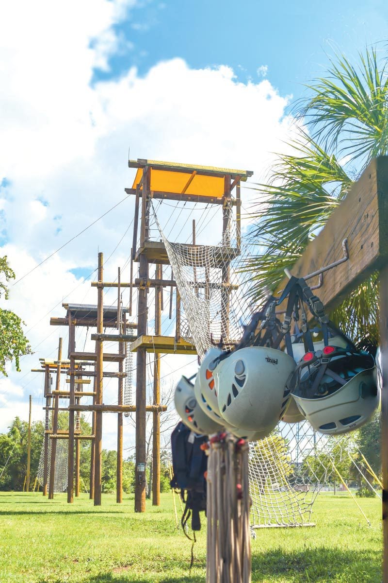 Helmets hang on the side of an empty high-elements climbing course made of ropes, wires, and wooden platforms and support beams. Helmets hang on the side of an empty high-elements climbing course made of ropes, wires, and wooden platforms and support beams.