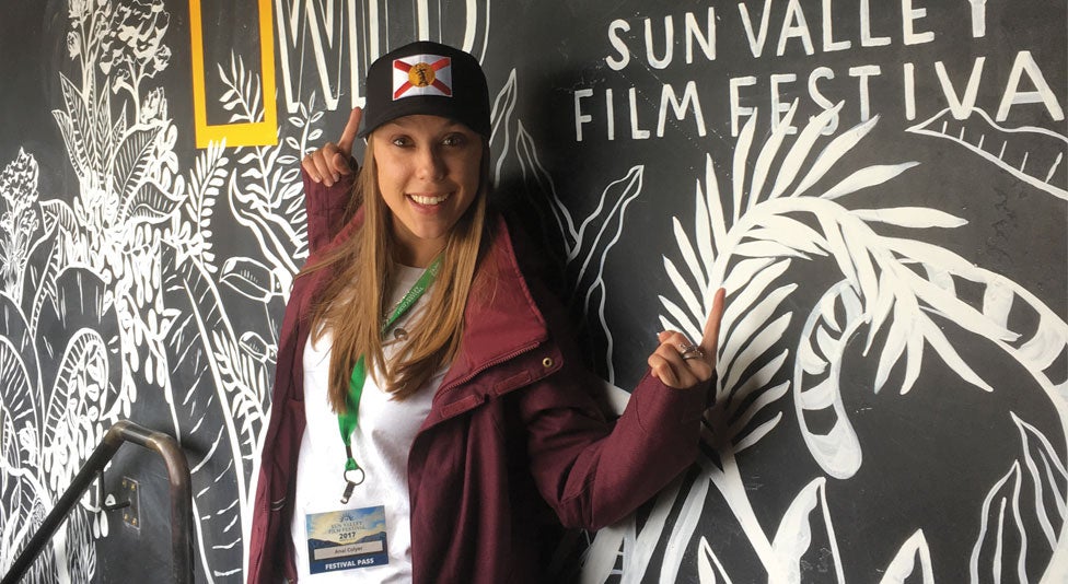 Anai Coyler, wearing a black baseball cap with the Florida flag, stands in front of a chalkboard illustration, pointing to two logos: one for Nat Geo Wild and one for Sun Valley Film Festival
