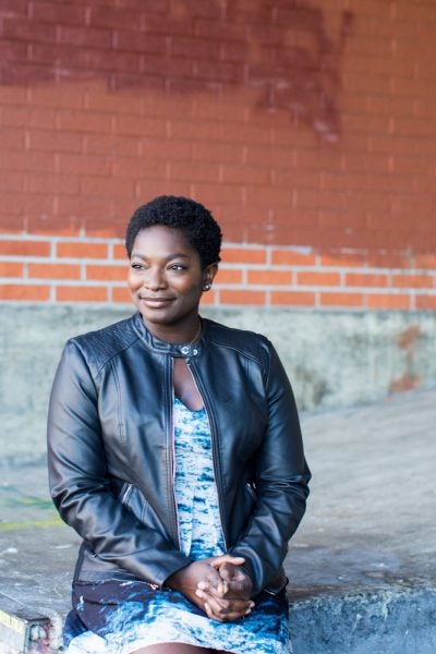 Melissa Amoi-Belinda Smith wearing a black leather jacket sits in front of a brick wall as she clasps her hands and looks off into the distance.