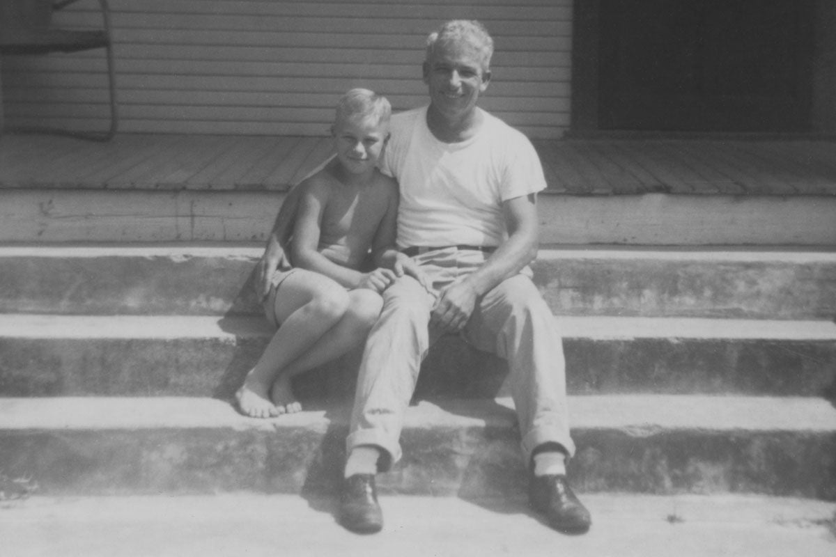 A black and white photo of a young, shirtless, white boy sitting next to an older white man, who has his arm around the younger boy, on the steps.