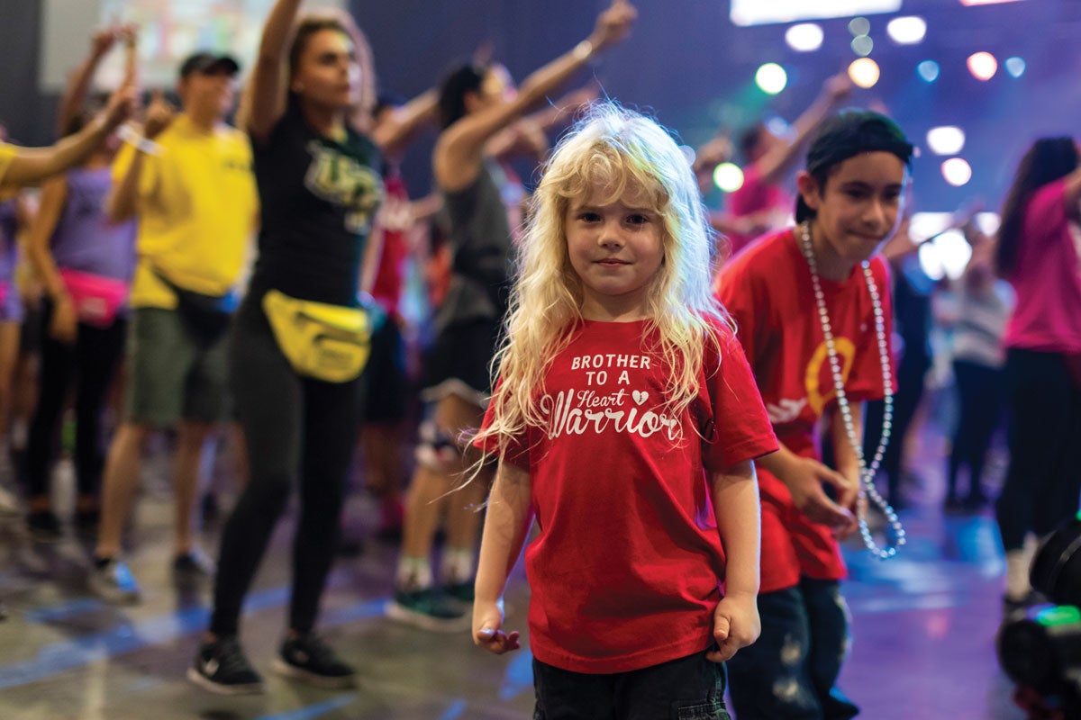A young child with long blonde hair smiles at the camera as a crowd of people dance behind her.