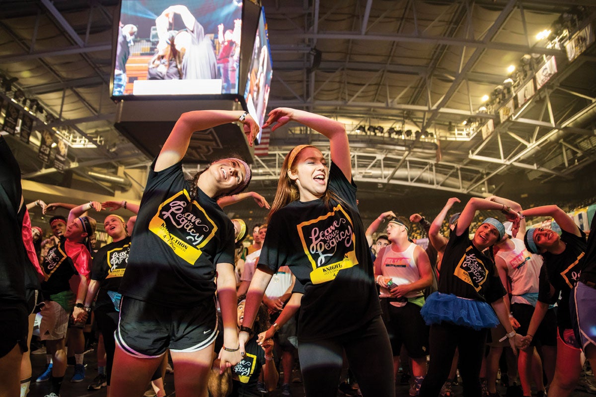 Two female students use their arms to form a heart while a crowd of people dance behind them in an arena.