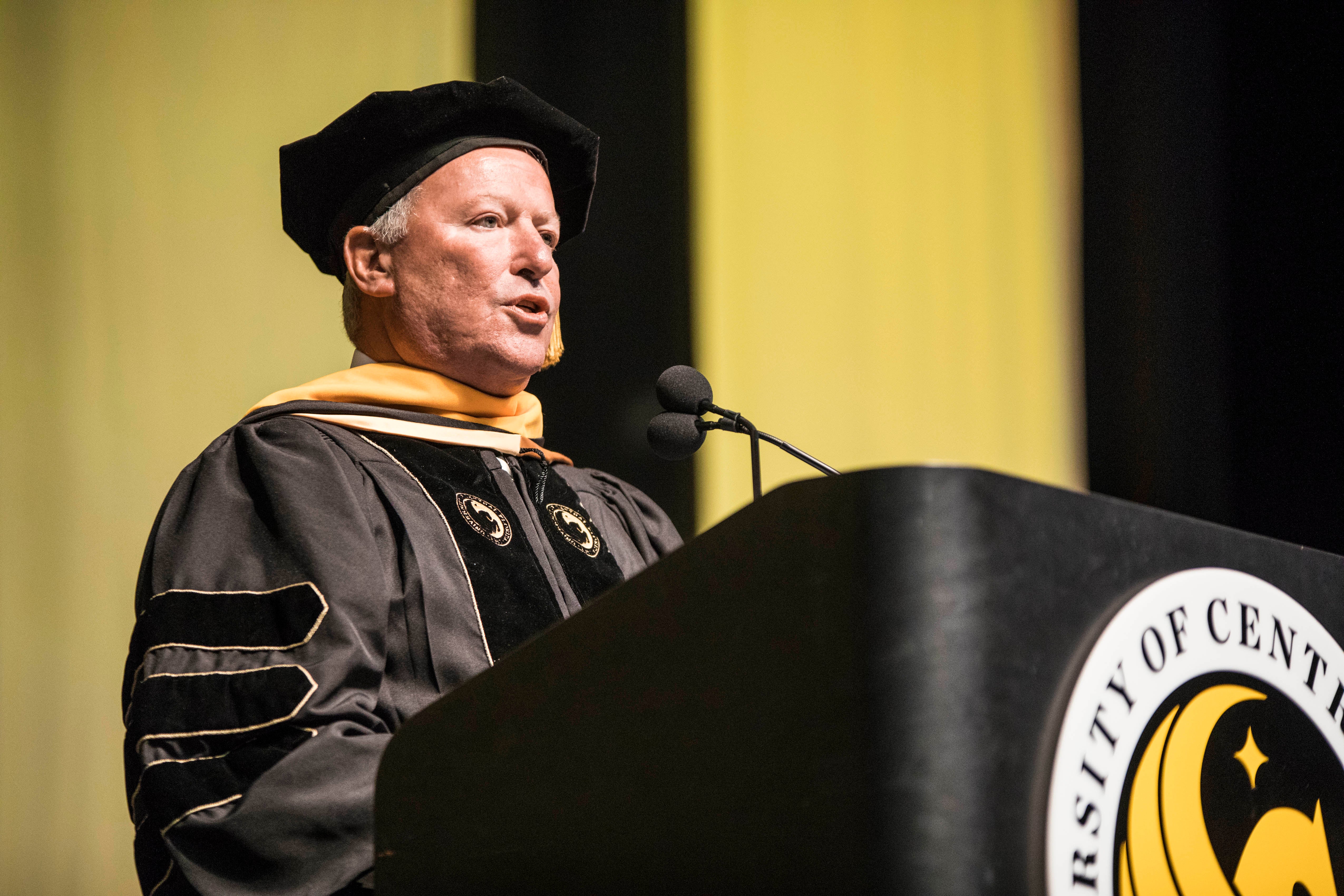 Orlando Mayor Buddy Dyer speaks at a podium while wearing a cap and gown.