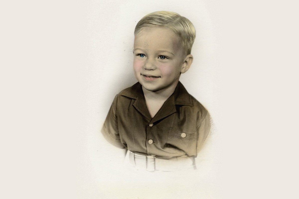 A colorized black and white photo of young white boy with blonde hair, blue eyes, and rosy cheeks wears a brown button-up shirt.