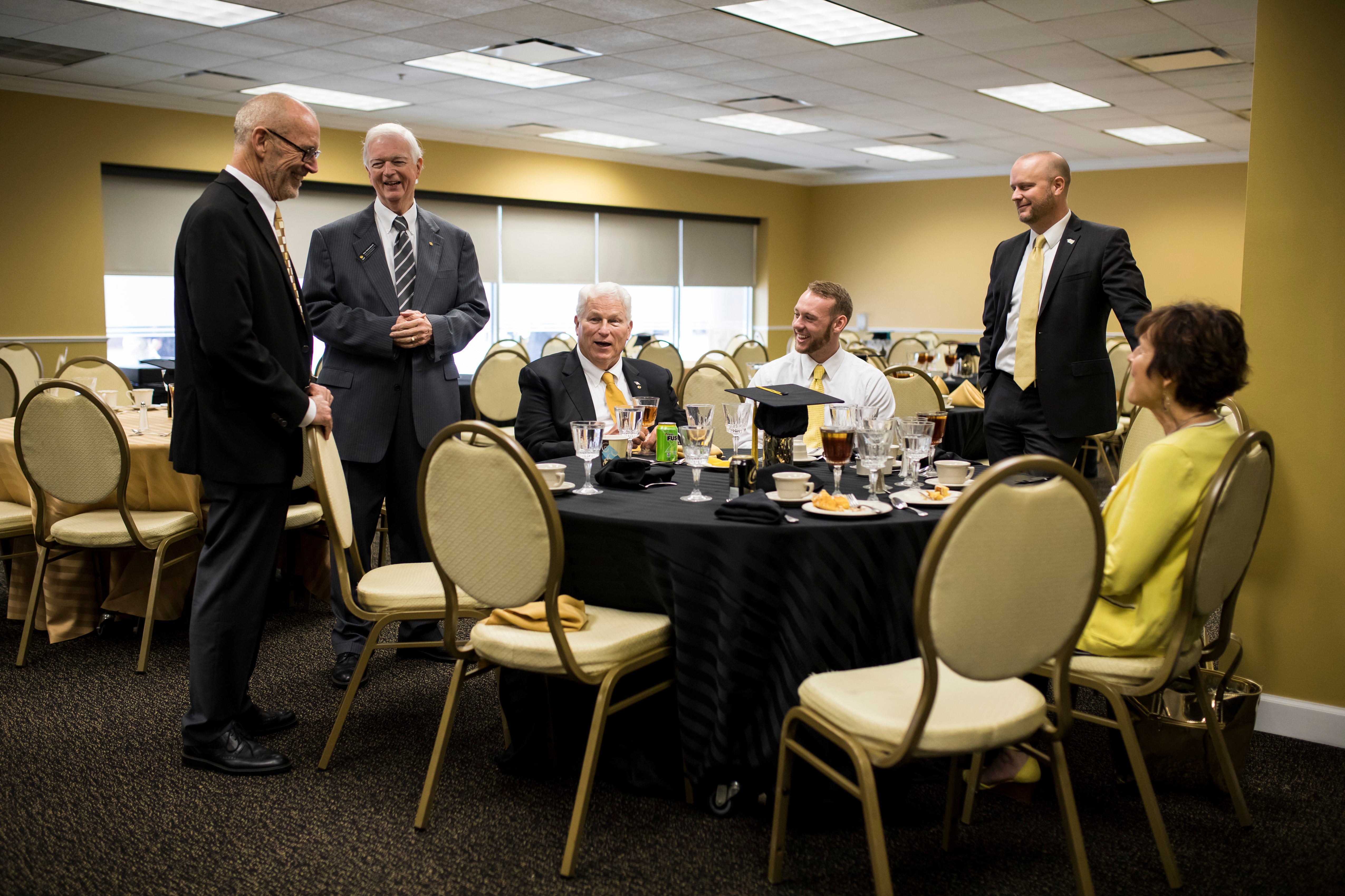 ohn C. Hitt sits at a table with a man and woman as three men stand around the group.