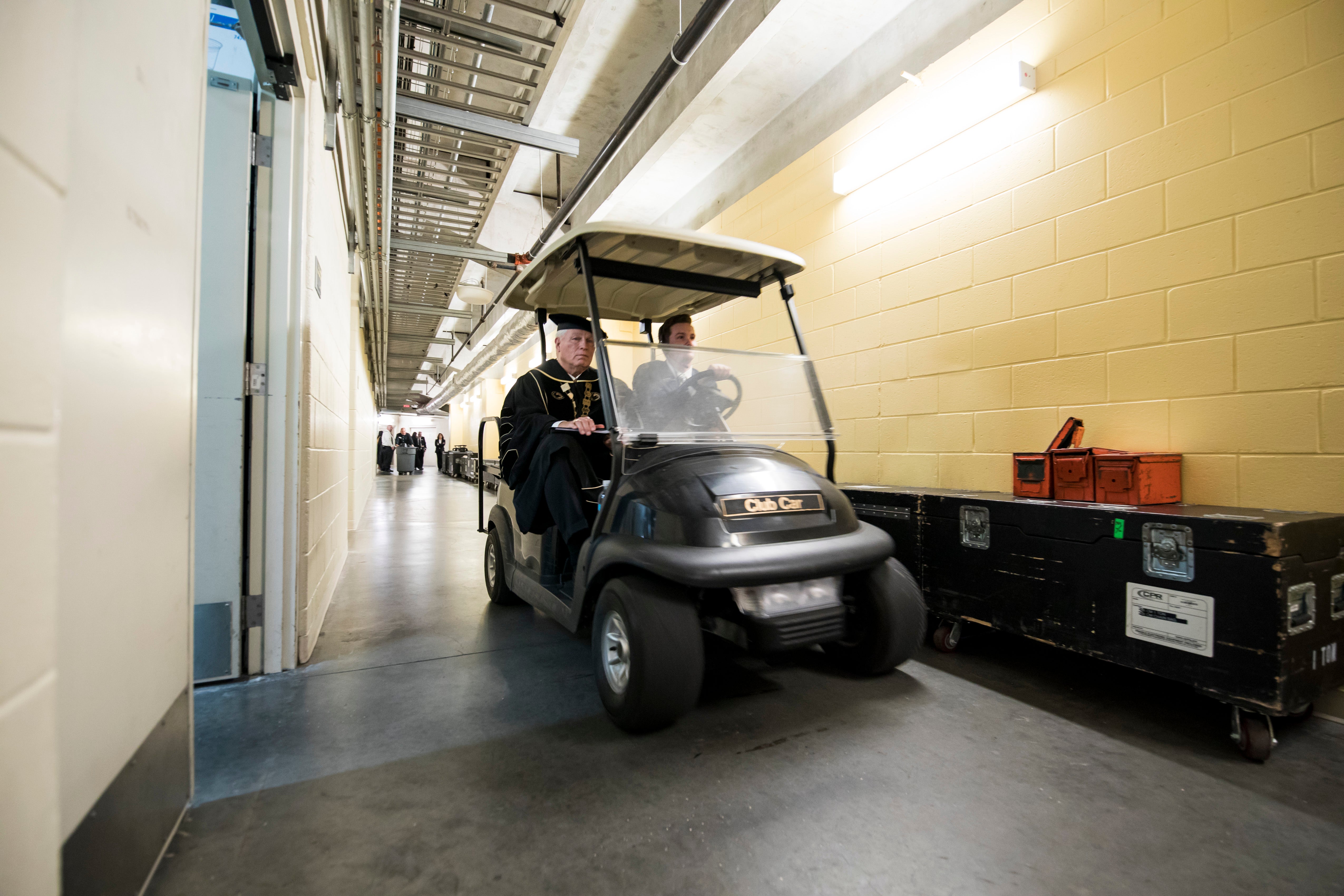 A man drives a golf cart through a hallway in CFE Arena as John C. Hitt wears a cap and gown and sits in the passenger seat .