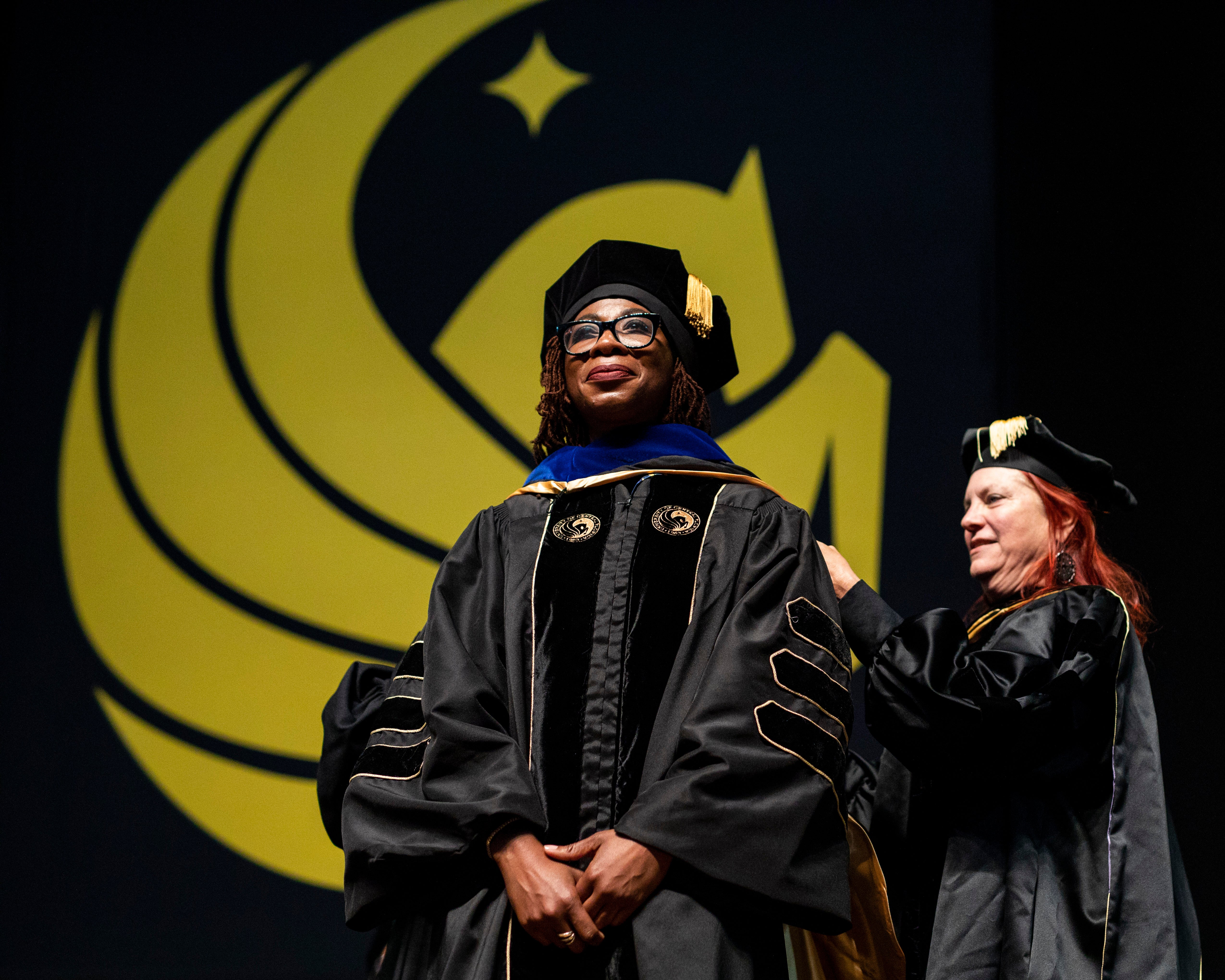 A female doctoral candidate waits on stage as a woman places a hood over her.