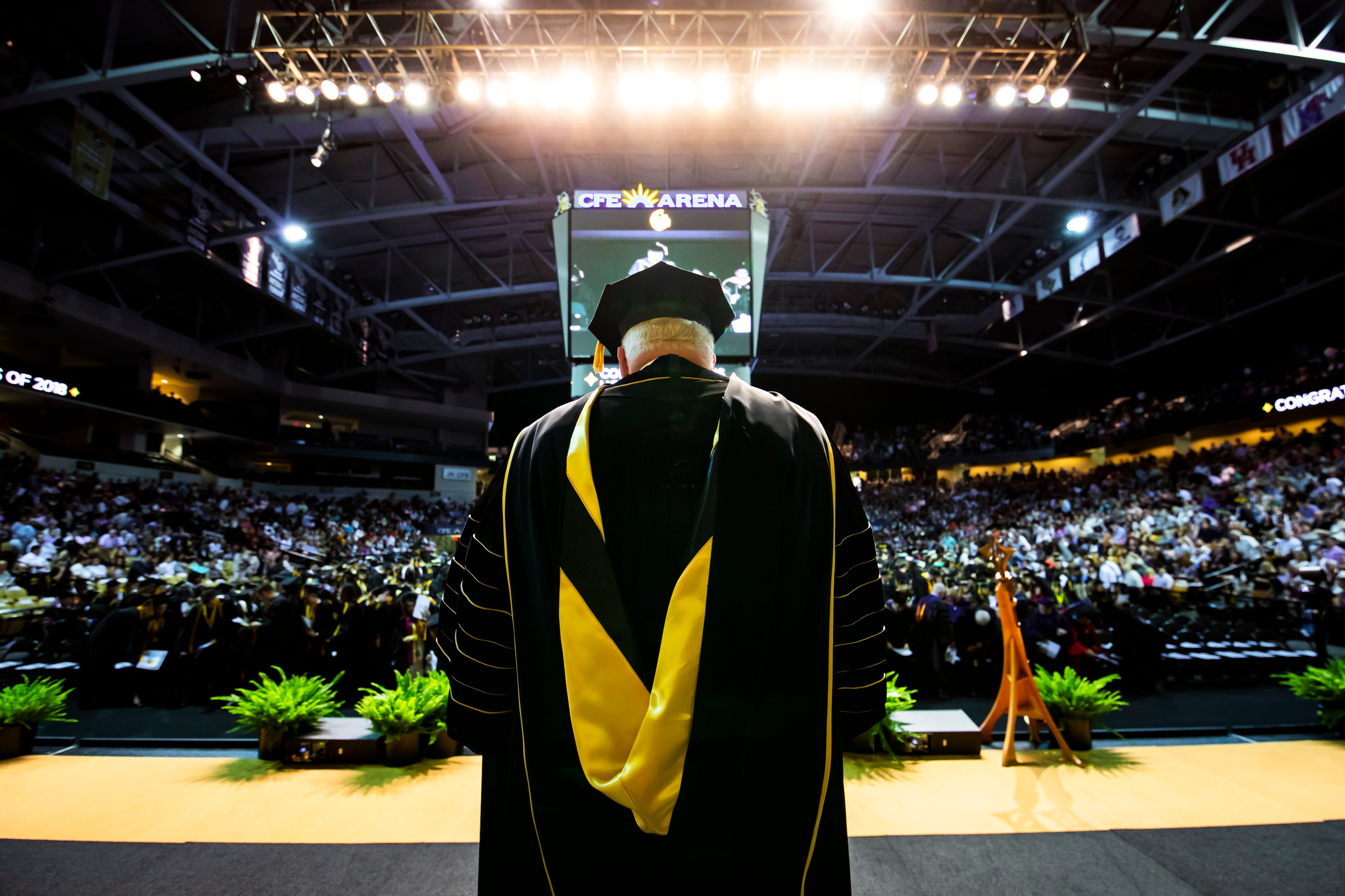 An image shows the back of John C. Hitt at a podium on stage as a crowded audience faces.