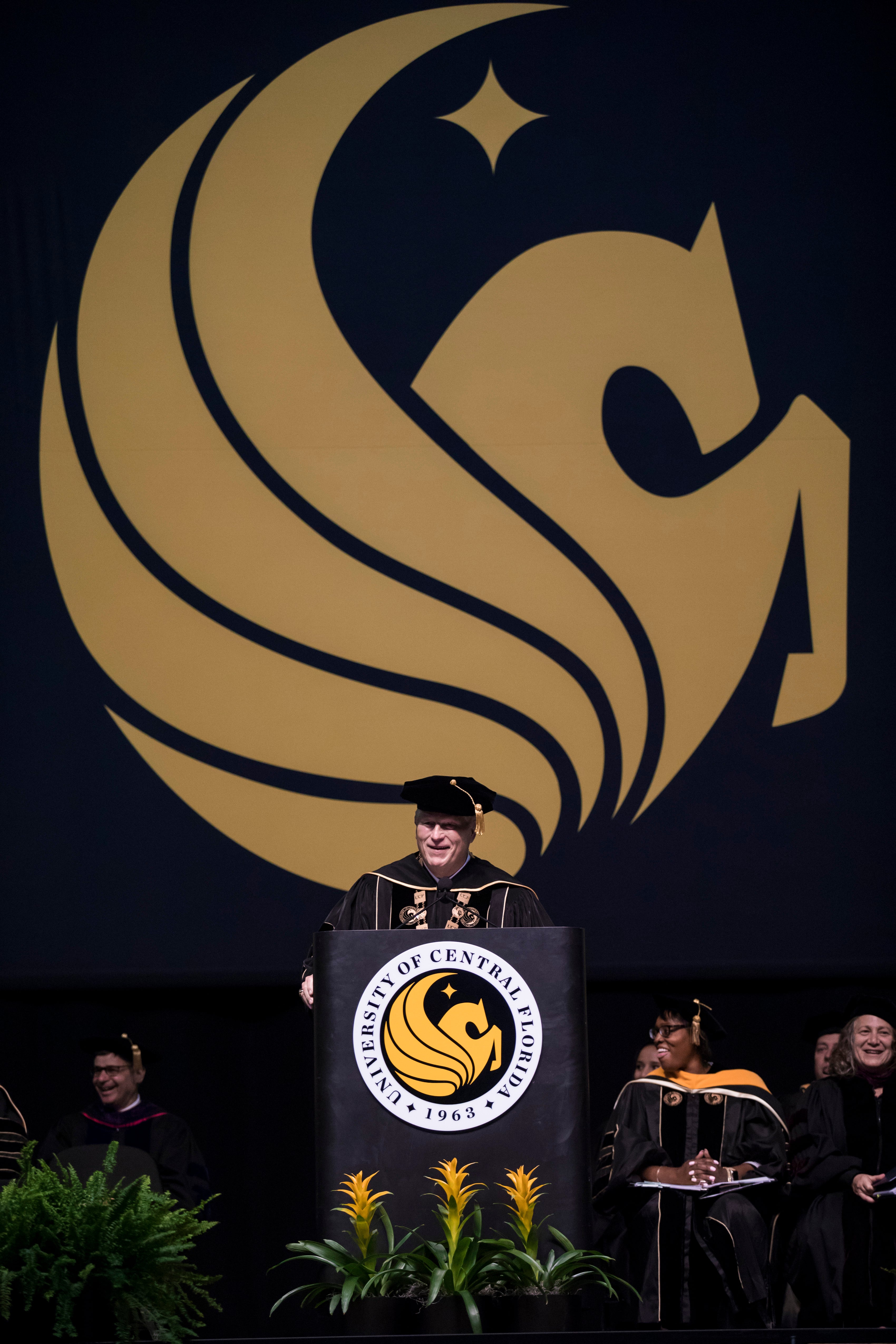 John C. Hitt speaks at a podium while wearing regalia as a yellow Pegasus symbol hangs behind him.