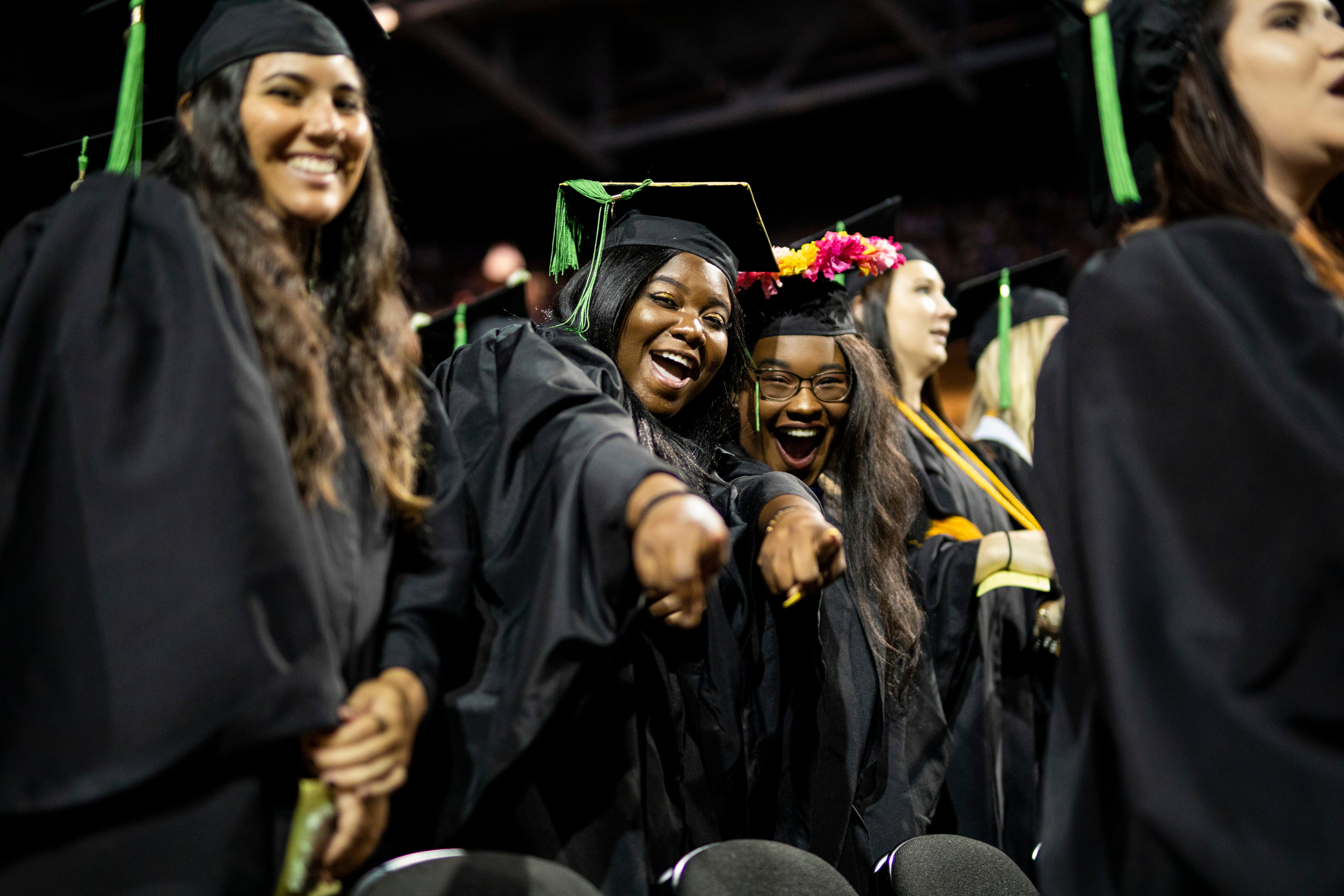 Two students wearing caps and gowns smile and point at the camera during a graduation ceremony.