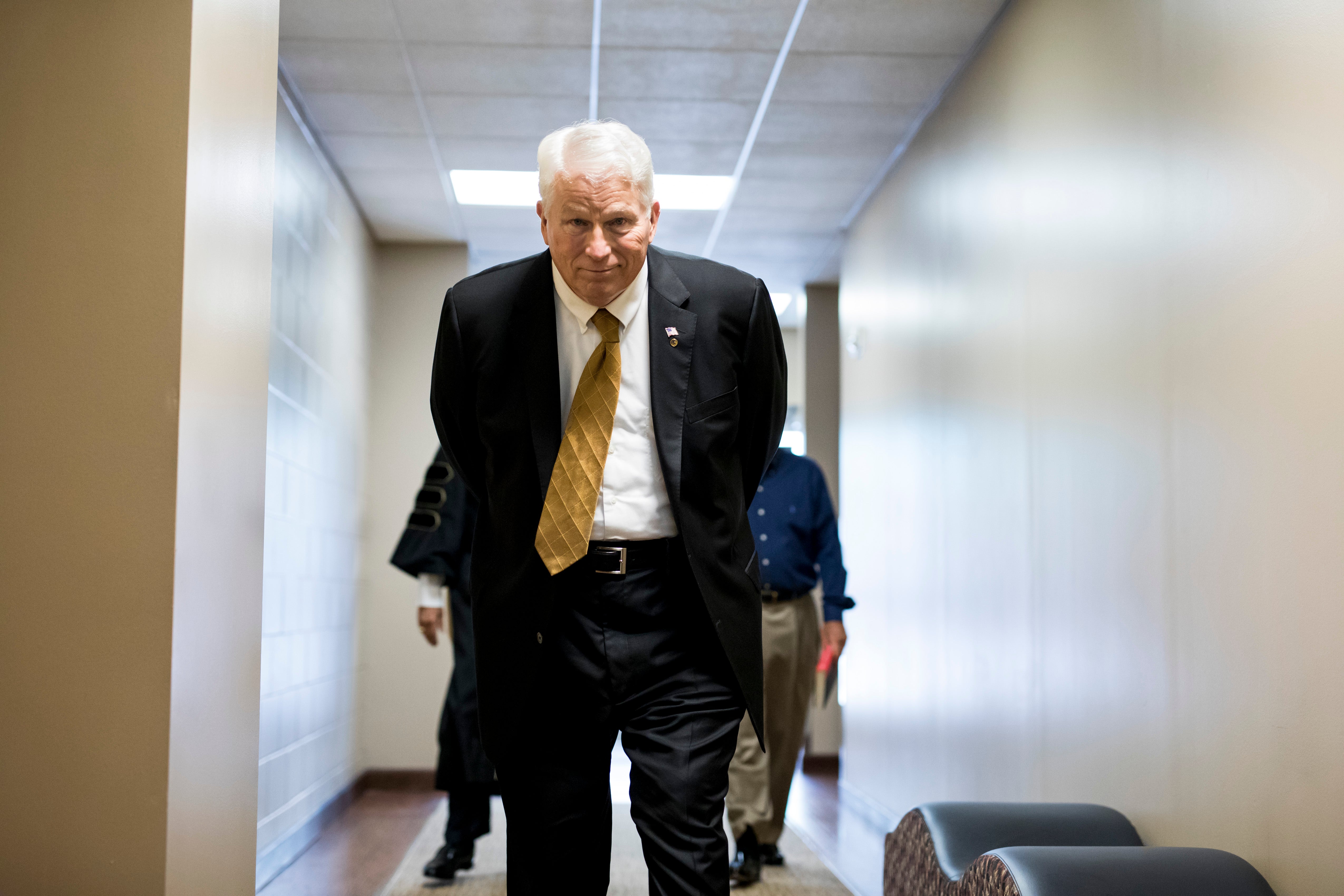 John C. Hitt wears a suit as he walks down a hallway with his hands behind his back.