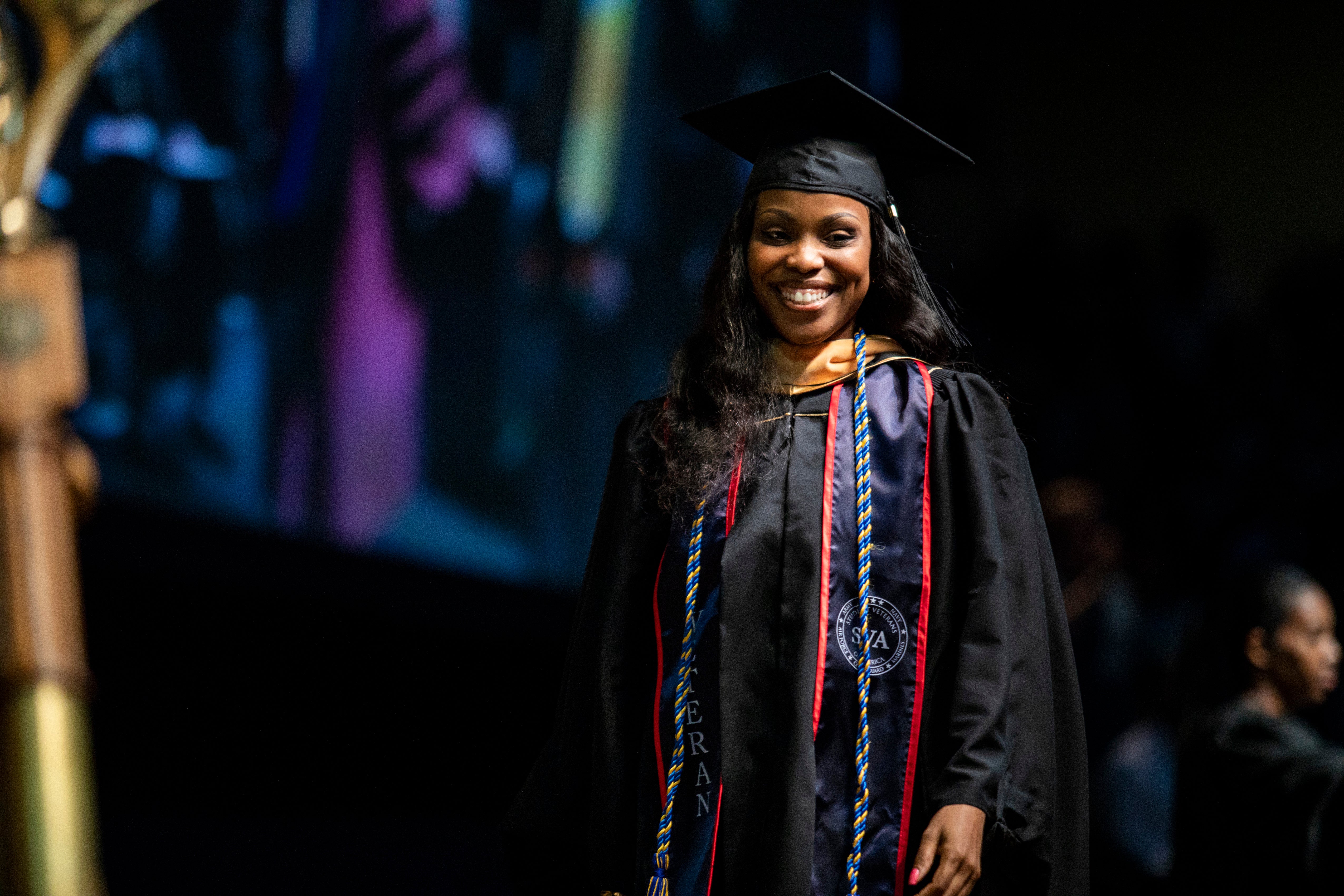 A female student smiles and wears a cap, gown and sash designating that she is a veteran as she crosses a stage.