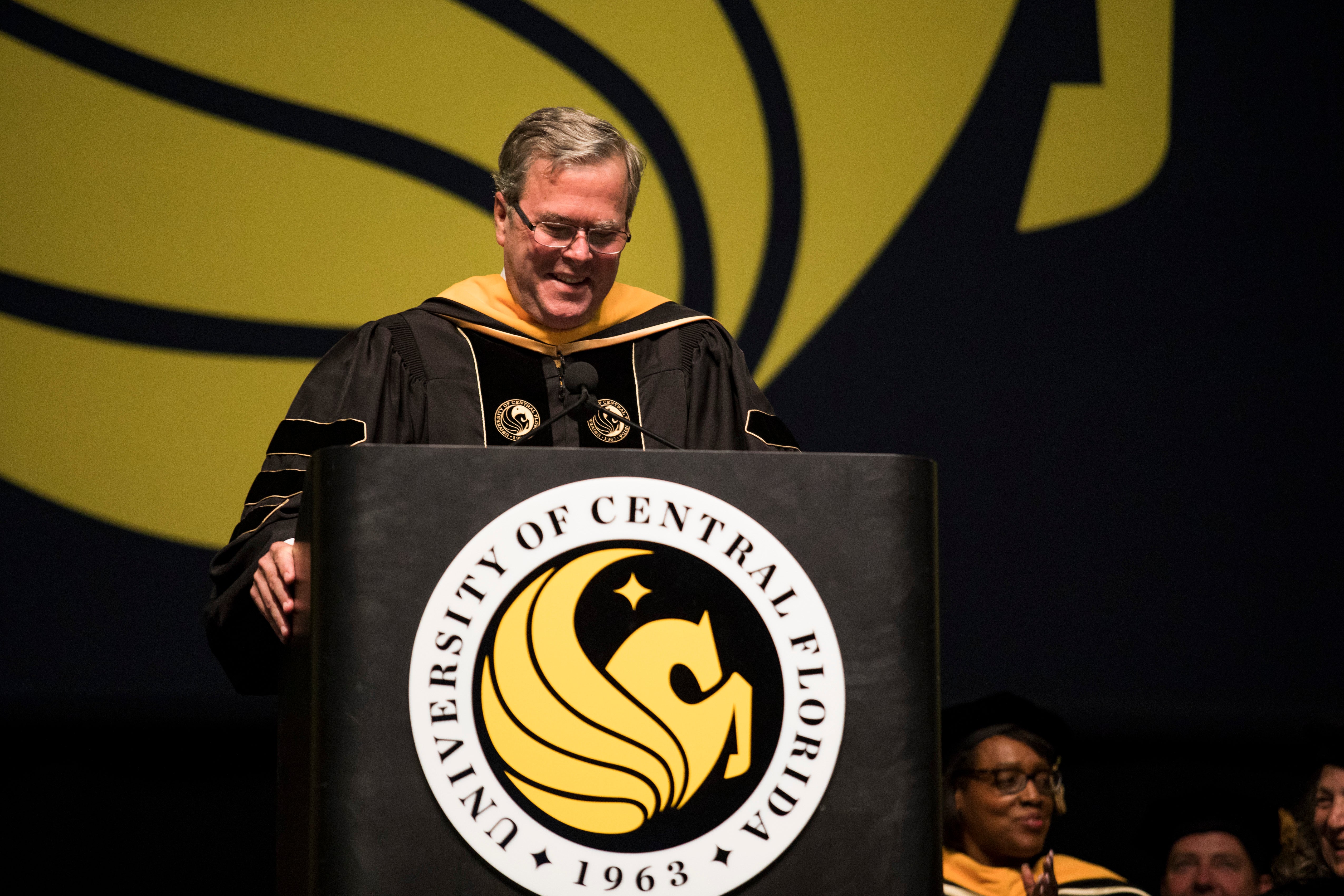 Former Florida Governor Jeb Bush wears a graduation gown as he stands behind a podium with the University of Central Florida Pegasus emblem on it.