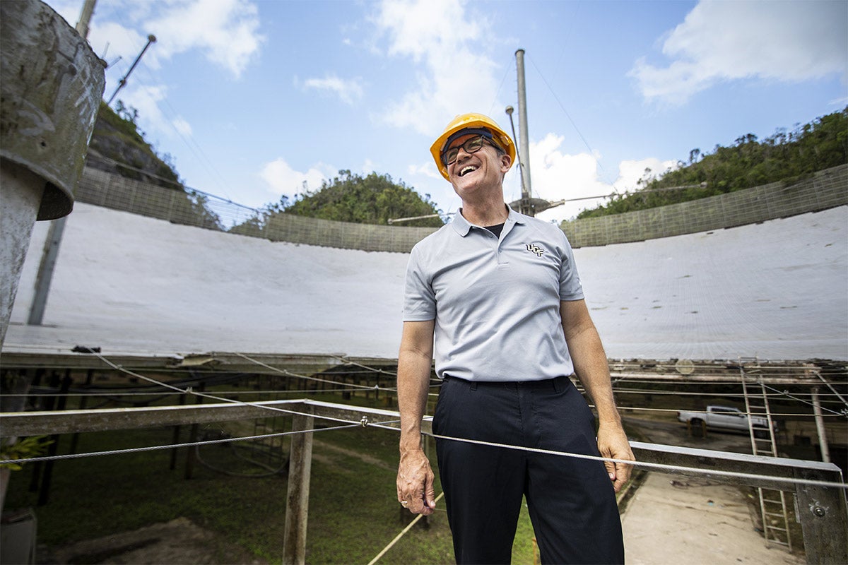 UCF President Dale Whittaker wearing yellow hard hat and glasses, grey polo, black pants, smiling near Arecibo.