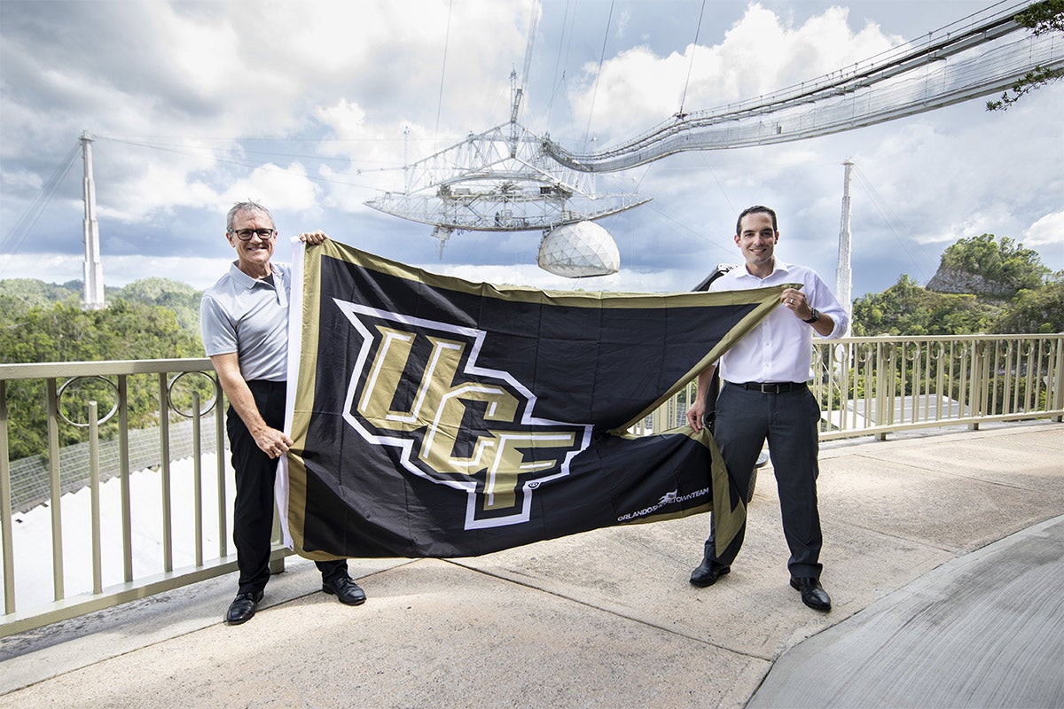 President Dale Whittaker and man holding up black and gold ucf flag at Arecibo. President Dale Whittaker and man holding up black and gold ucf flag at Arecibo.