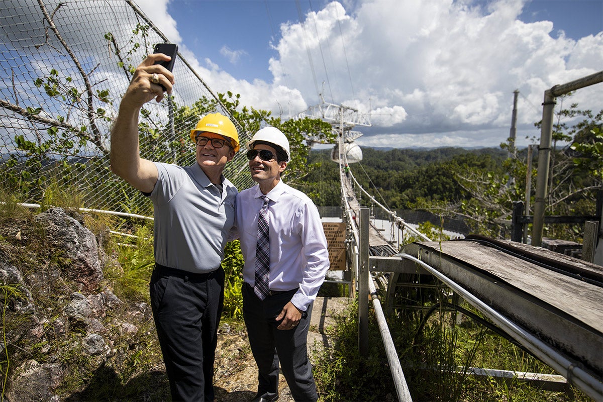 President Dale Whittaker wearing a grey polo, yellow hard hat takes a selfie with man wearing purple shirt, plaid tie, sunglasses and a white hard hat in Arecibo complex.