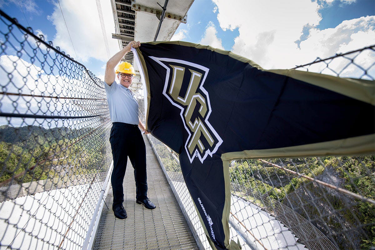 A man in a polo and hardhat holds up a giant black UCF flag.