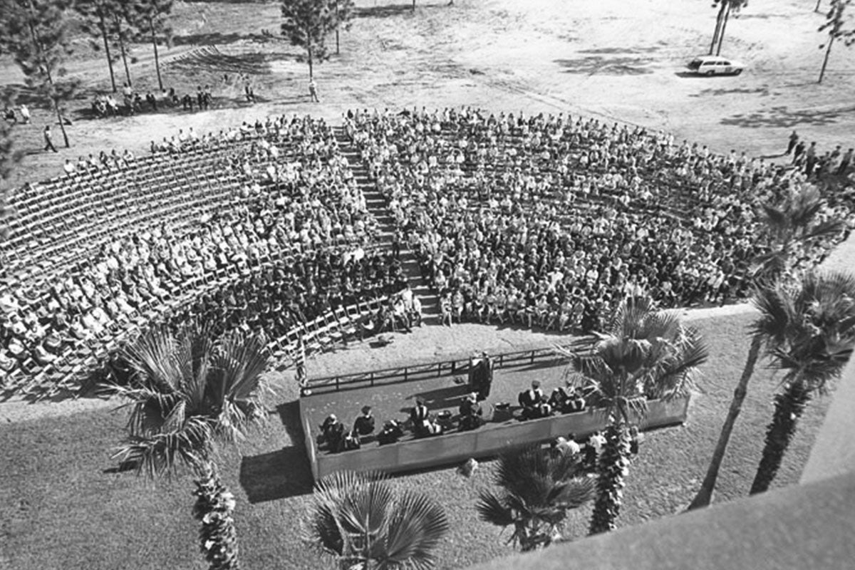 A black and white overhead shot shows a large crowd outside while a man speaks at a podium.