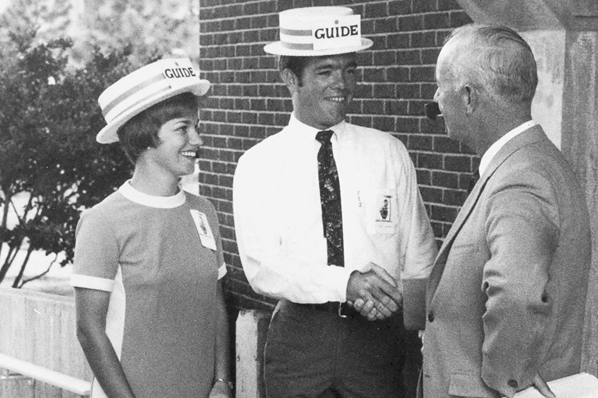 A black and white image shows two students wearing hats while one shakes hands with a man in a suit.