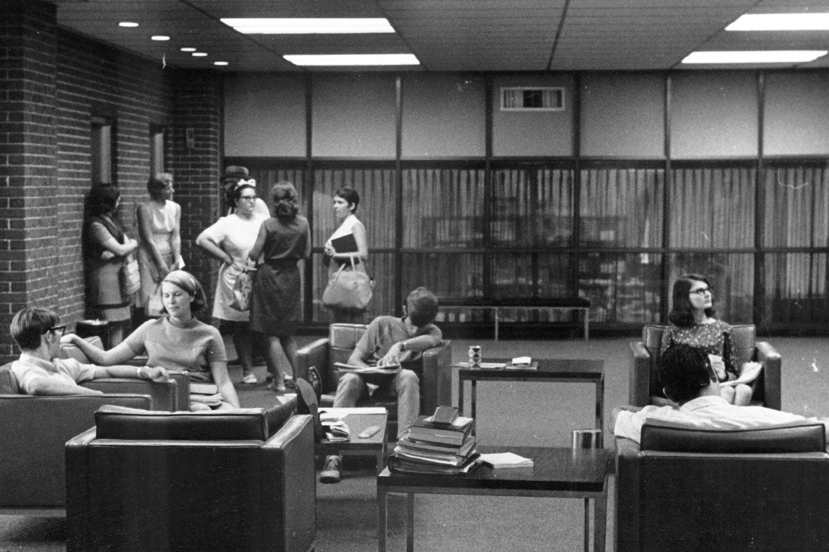 A black and white image shows a group of students standing at the back of a room, while others sit in chairs with books.