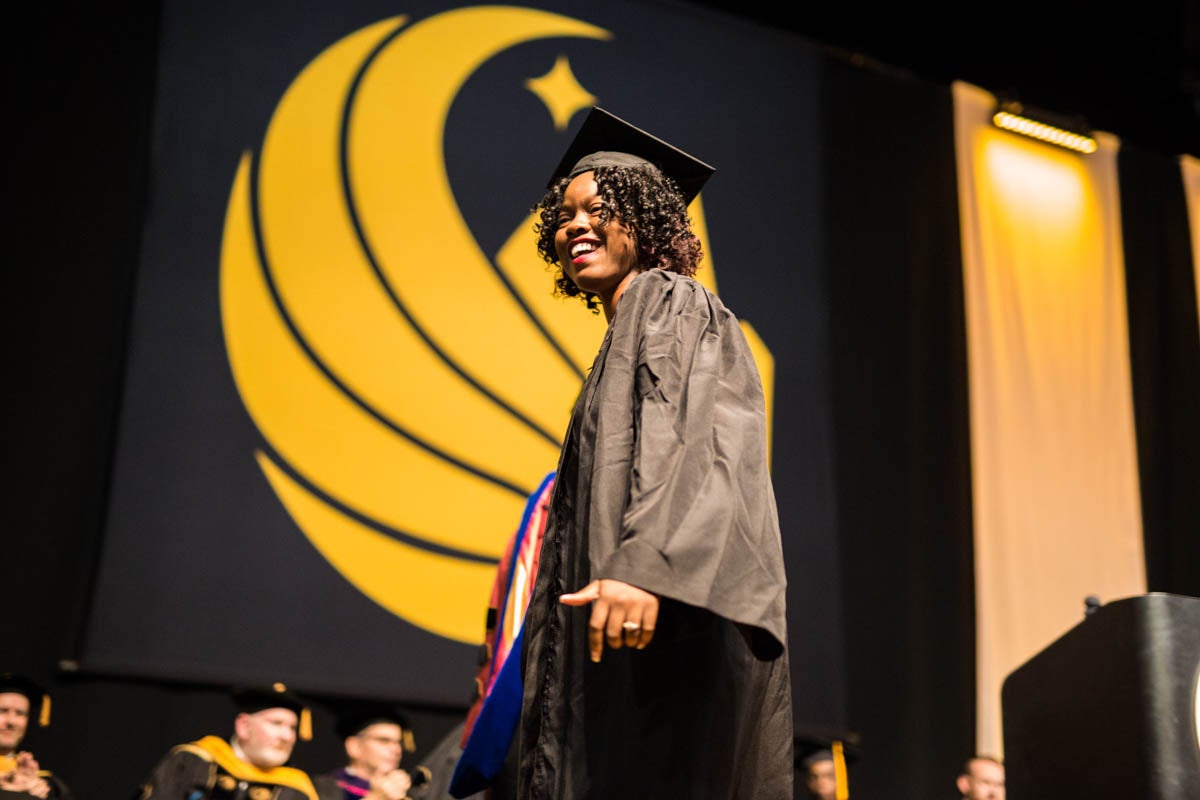 A woman in a cap and gown walks across the stage with a yellow pegasus symbol in the background.