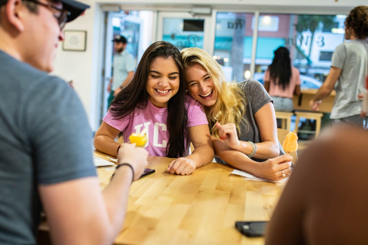 Two female students laugh and lean on each other while they sit at a table and eat popsicles.