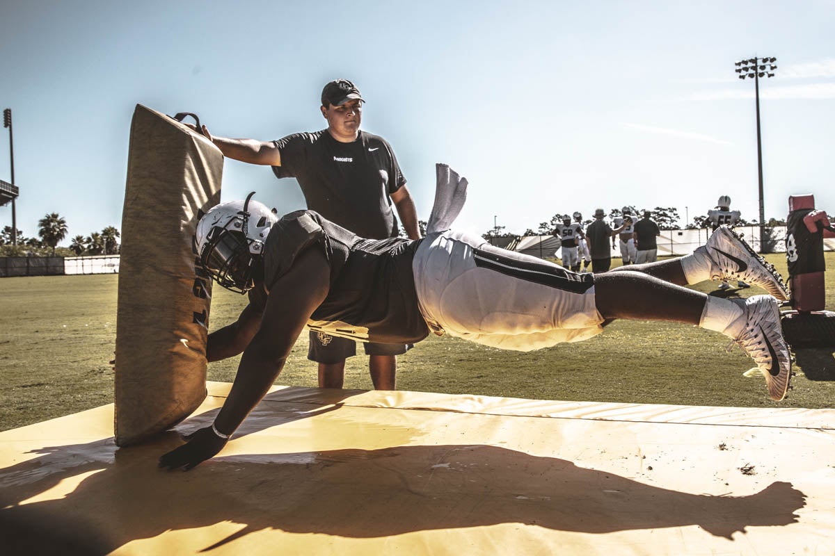 A football player hovers over a mat as he crashes into a talking pad that a man is holding.